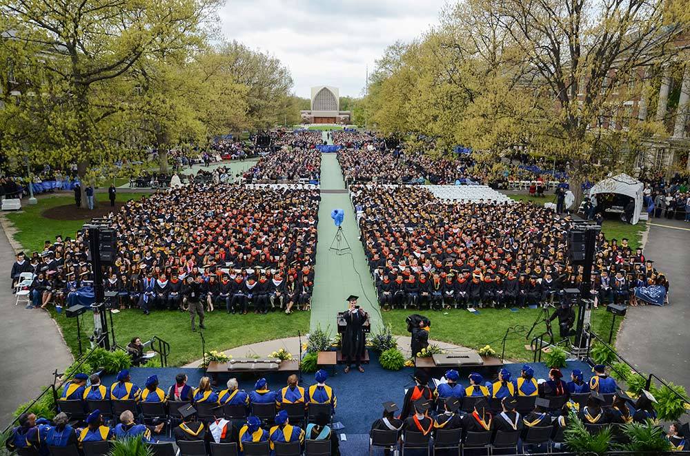 Large outdoor college commencement crowd seated on a campus lawn under the sun