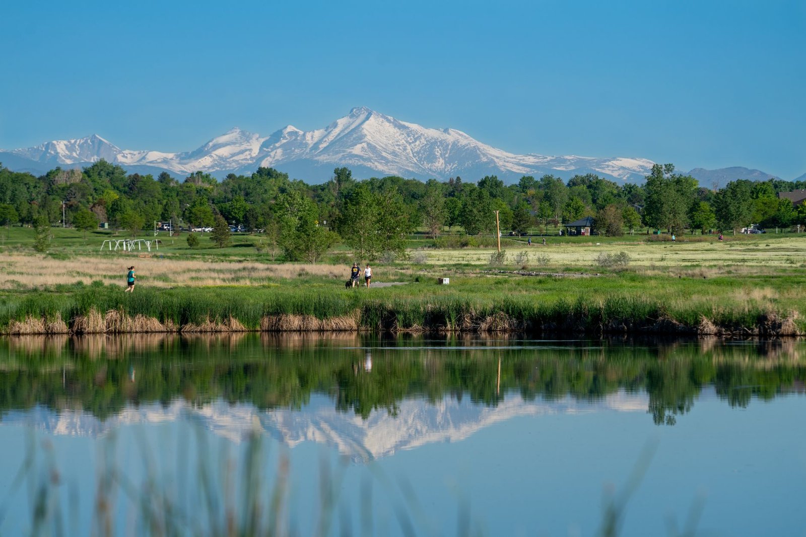 Walkers and a runner on a park trail beside a reflecting pond near the mountains