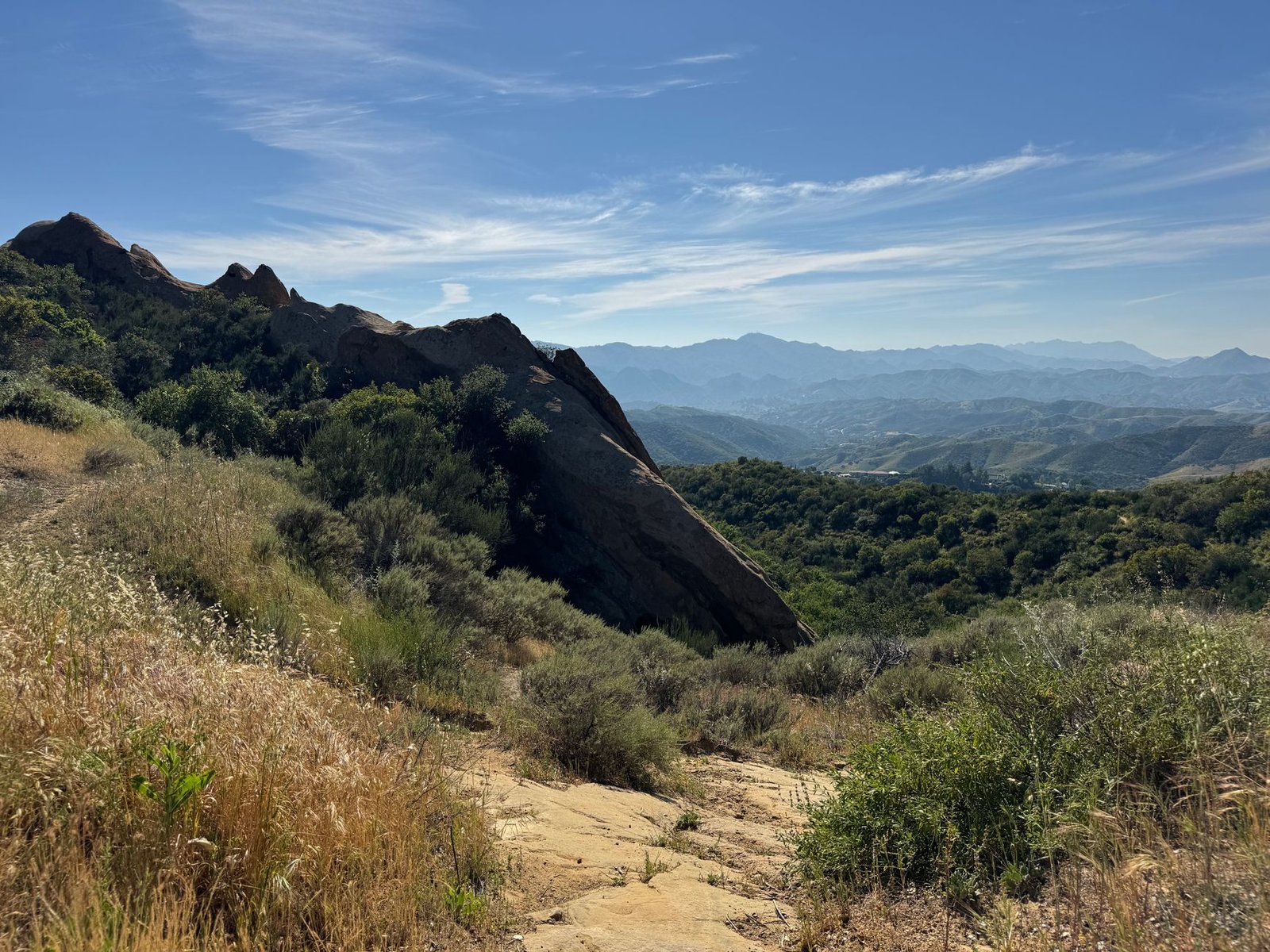 A rocky hillside trail through chaparral with mountains in the distance