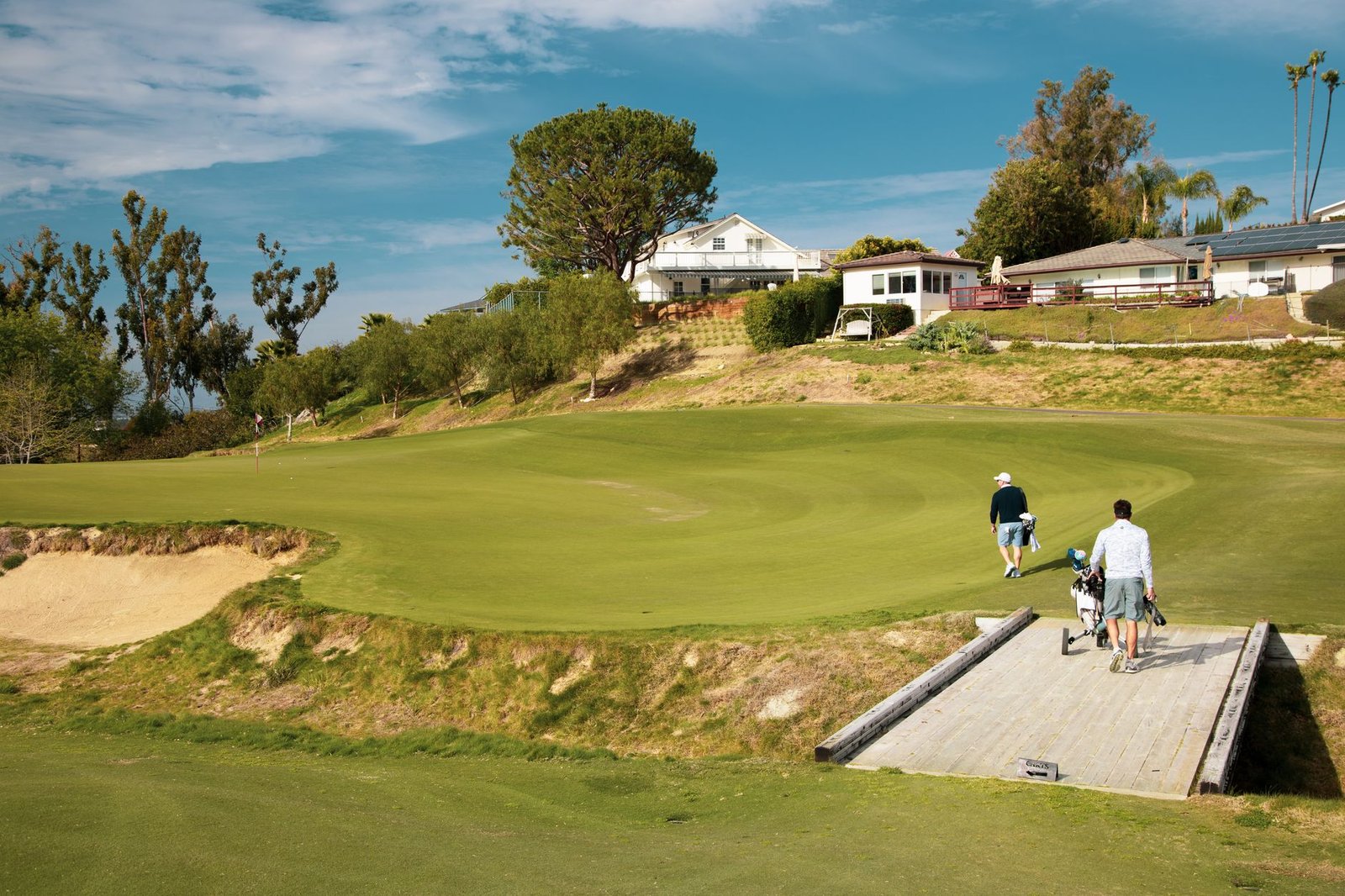 Two golfers walking up to a green on a hillside golf course with homes above