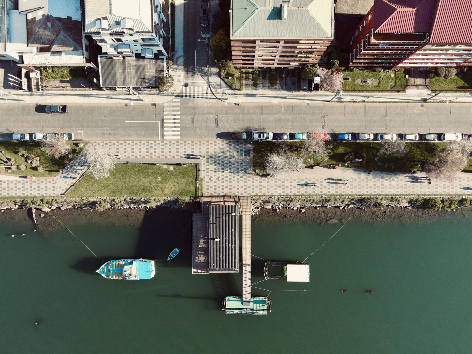 An aerial view of a riverfront promenade with parked cars, benches and moored boats